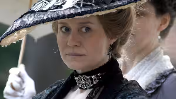 A woman in a black dress and feathered hat holds a parasol