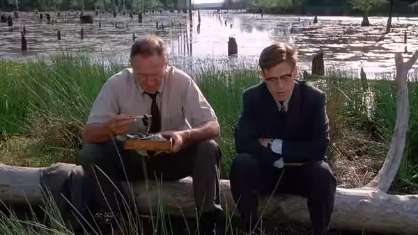Rupert Anderson (Gene Hackman) eats from a box while Alan Ward (Willem Dafoe) sits nearby on a log