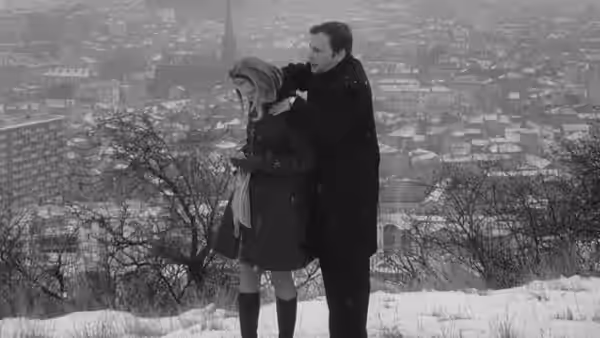 A man holds a woman close on a snowy street beneath bare trees