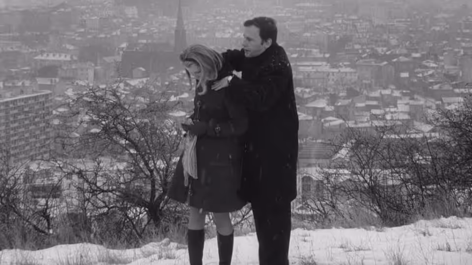 A man holds a woman close on a snowy street beneath bare trees