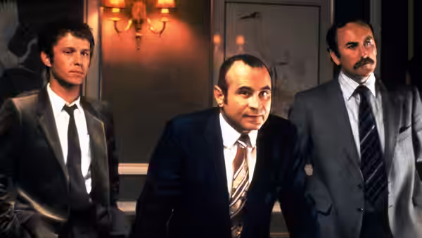 Harold Shand (Bob Hoskins) standing between two men in dark suits indoors