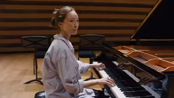 woman playing grand piano turning to look back with another musician's hands on keys