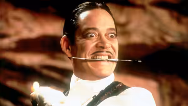 Man with slicked hair and thin mustache holding a candle with a needle through his head