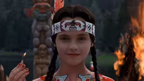 Wednesday Addams in Native American costume holding a lit match by a fire