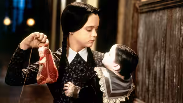 Wednesday Addams holding raw meat on a string while a baby watches