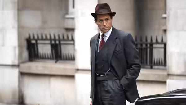 Jeremy Thorpe (Hugh Grant) standing outside a stone building in a suit and hat