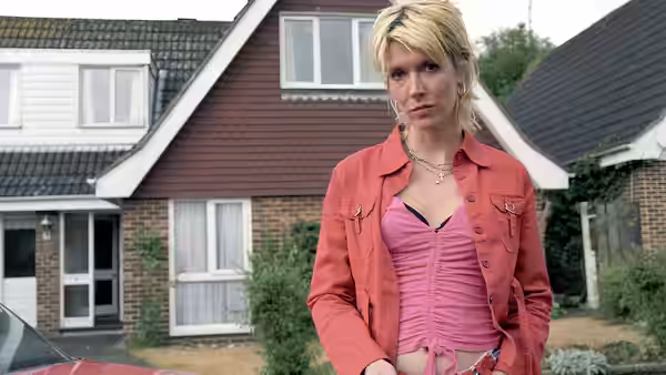 Jill Tyrrell (Julia Davis) stands outside suburban houses wearing a red jacket