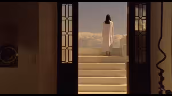 A woman in a white dress stands beyond an open doorway at the top of a staircase