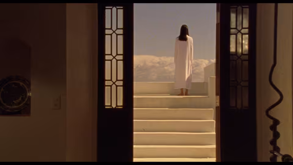 A woman in a white dress stands beyond an open doorway at the top of a staircase