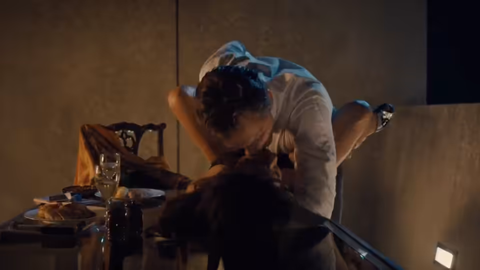 A man leans over another person beside a dining table