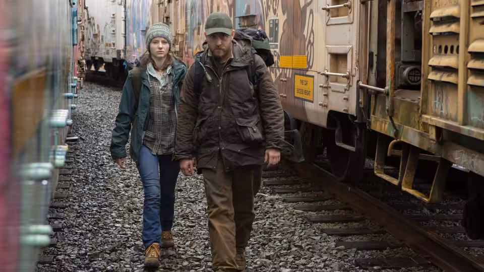 Tom (Thomasin McKenzie) and Will (Ben Foster) walking hand in hand between freight trains