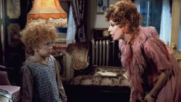 Miss Hannigan (Carol Burnett) bends to speak with Annie (Aileen Quinn) in a parlor
