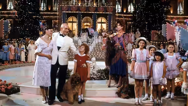 Daddy Warbucks (Albert Finney) stands with Annie (Aileen Quinn) and Miss Hannigan (Carol Burnett) on a lit stage