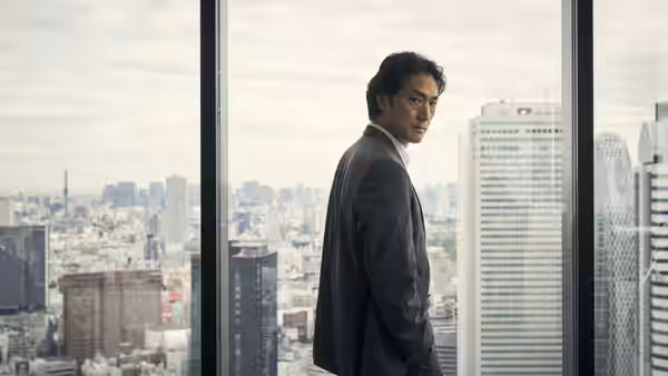 Man in a grey suit looks back from a high-rise window over a city