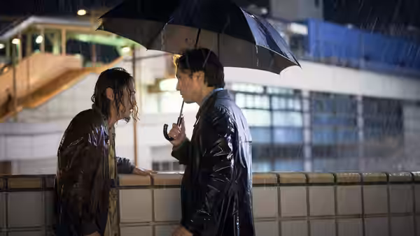 Two men stand close under a black umbrella in heavy rain at night