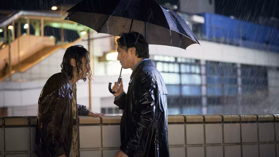 Two men stand close under a black umbrella in heavy rain at night