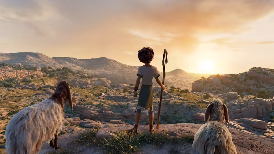 Young shepherd boy with staff stands between two goats on rocky hillside at sunset