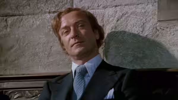 Milo Tindle (Michael Caine) seated against a stone wall in a suit