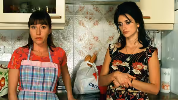 Two women stand in a kitchen, one in a plaid apron, the other with arms crossed