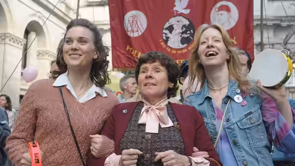 Three women stand together at an outdoor rally holding a tambourine