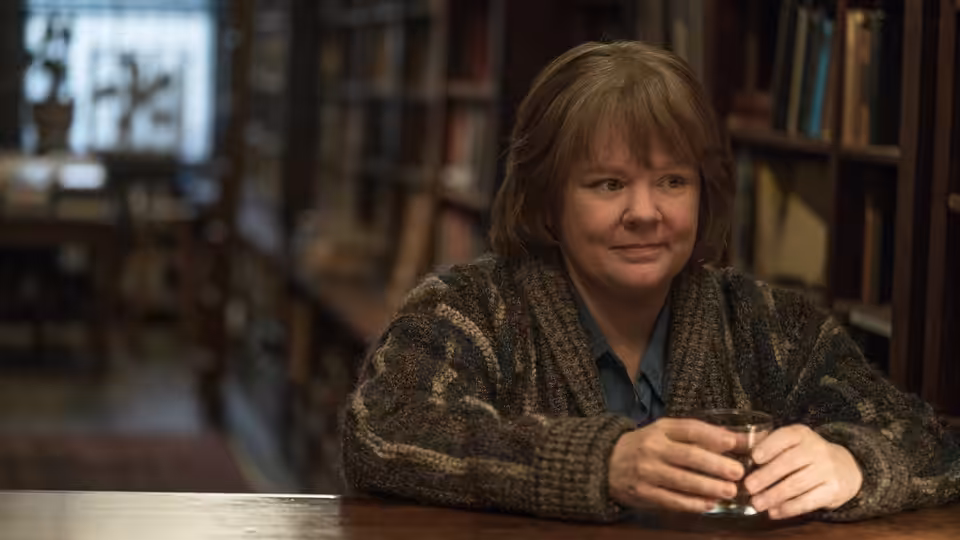 Lee Israel (Melissa McCarthy) holding a glass at a library table