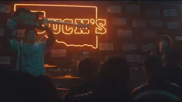 Musician raises a guitar on stage beside a glowing neon sign with another performer