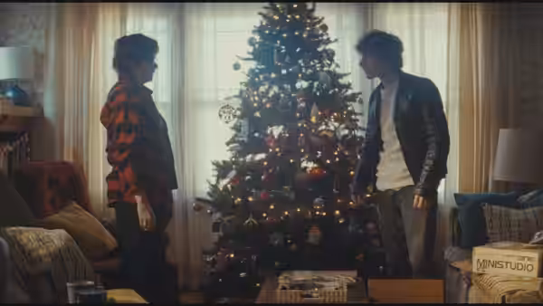 Two young men face each other beside a lit Christmas tree in a living room