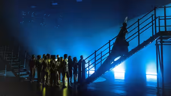 A robed performer descends stairs as a group watches from the stage floor