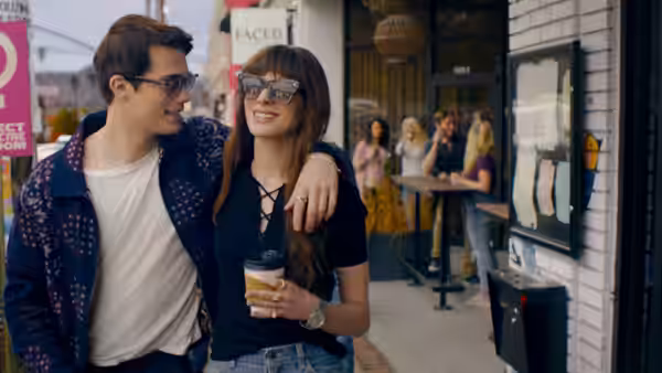 Hayes (Nicholas Galitzine) with arm around Solène (Anne Hathaway) outside a cafe on a city sidewalk