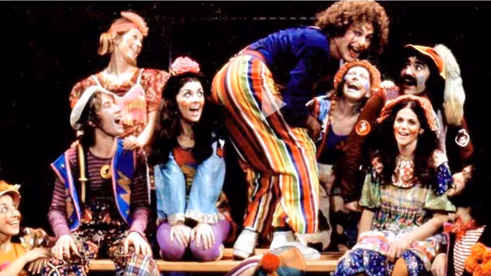 Performers in colorful costumes laugh and lean together on stage
