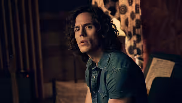 A curly-haired musician stands beside recording equipment in a dim studio