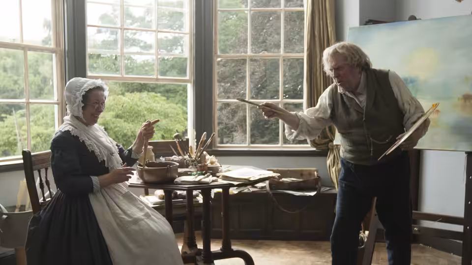 JMW Turner (Timothy Spall) gestures with a brush beside a seated woman in his studio