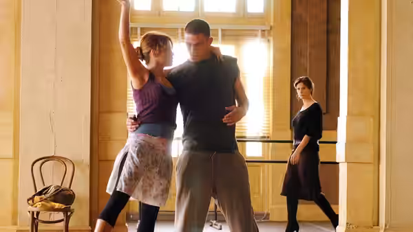 a couple dances closely in a sunlit studio while another woman watches
