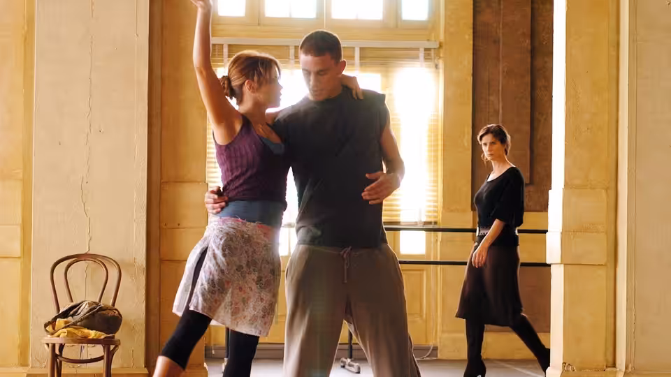 a couple dances closely in a sunlit studio while another woman watches