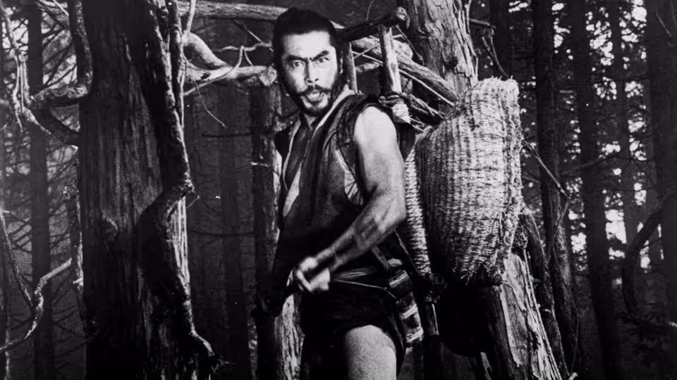 Tajômaru (Toshirō Mifune) standing among trees with a woven basket on his back