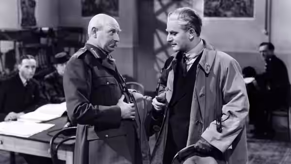 Two men in coats conversing in an office with others seated nearby