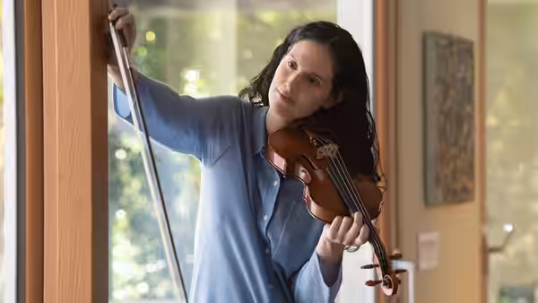 Nadia Hynes (Eva Victor) holding a violin by a window
