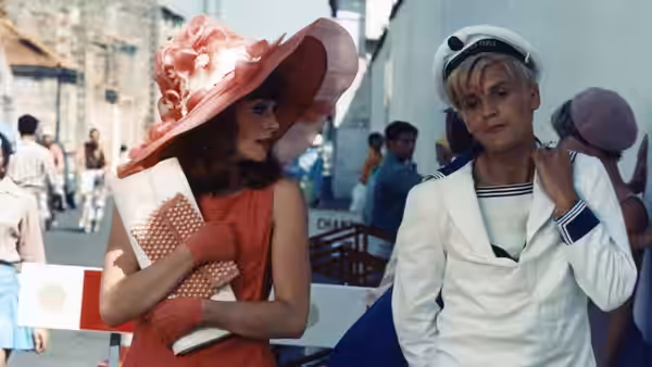 A woman in a broad pink hat stands beside a sailor in a white uniform