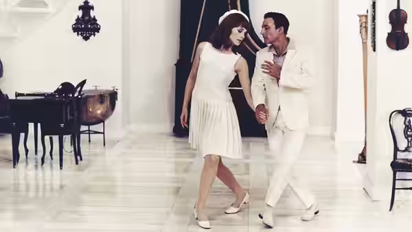 A woman in a white dress dances with a man in a white suit inside a music room