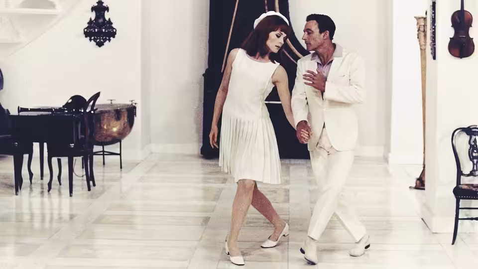 A woman in a white dress dances with a man in a white suit inside a music room