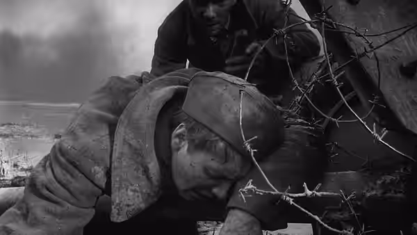 A wounded soldier lies beneath a blanket beside barbed wire