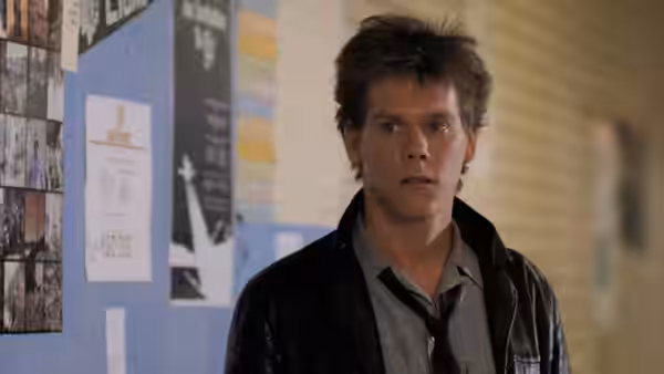 Ren (Kevin Bacon) standing in a school hallway with posters on the wall