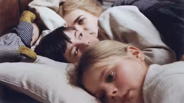 Three children lie together under blankets resting their heads on pillows