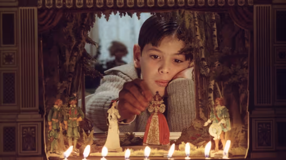 Boy in a knit sweater points at a figure in a candlelit puppet theater