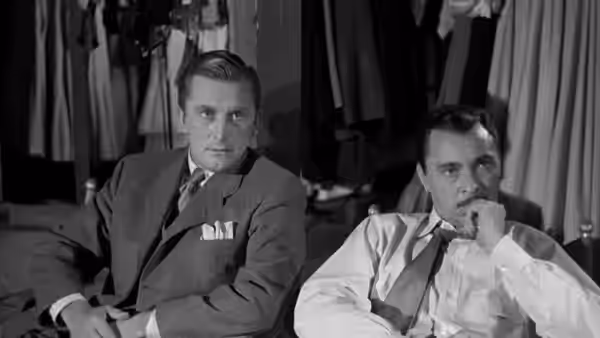 Two suited men sit pensively in a room lined with hanging clothes