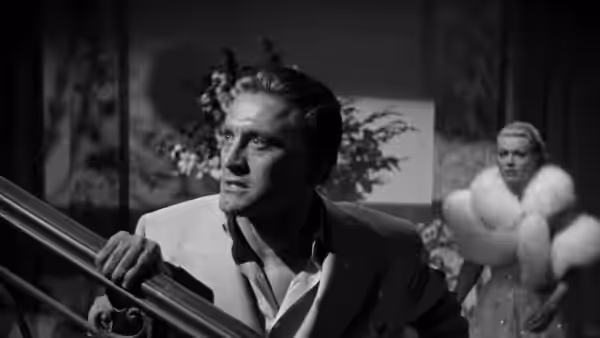 Jonathan Shields (Kirk Douglas) grips a staircase railing as a fur-clad woman watches