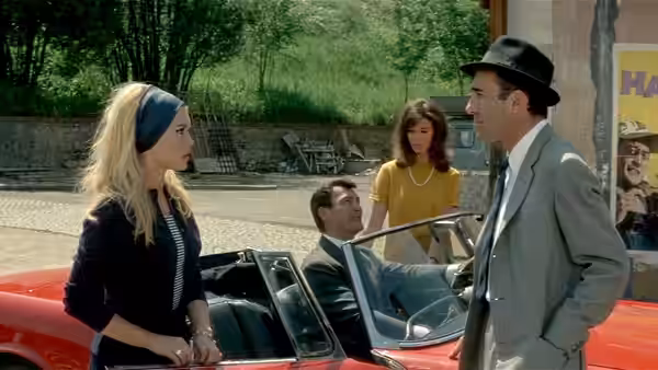 Paul Javal (Michel Piccoli) talks with Camille (Brigitte Bardot) beside a red convertible