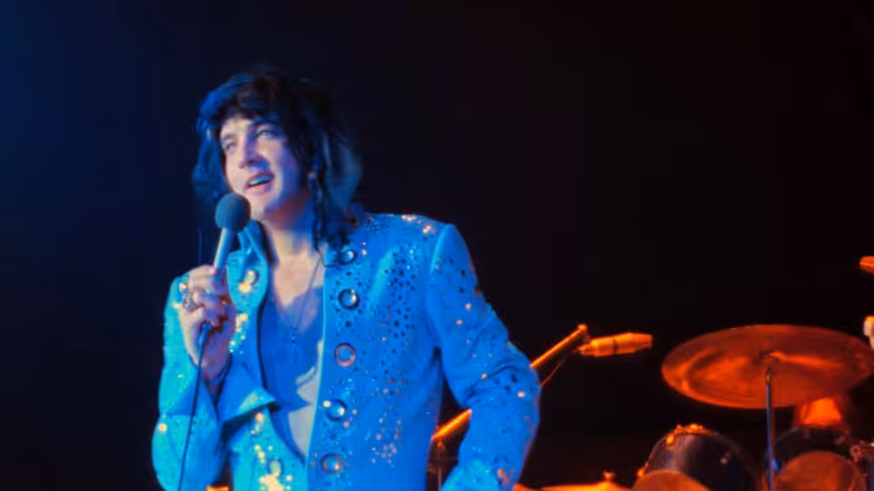 Elvis Presley singing into a microphone on a dark stage with drums behind