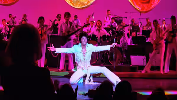 Elvis Presley kneeling on stage with arms outstretched in front of an orchestra