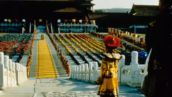 Rows of officials in colorful robes kneel on stone steps before a distant throne